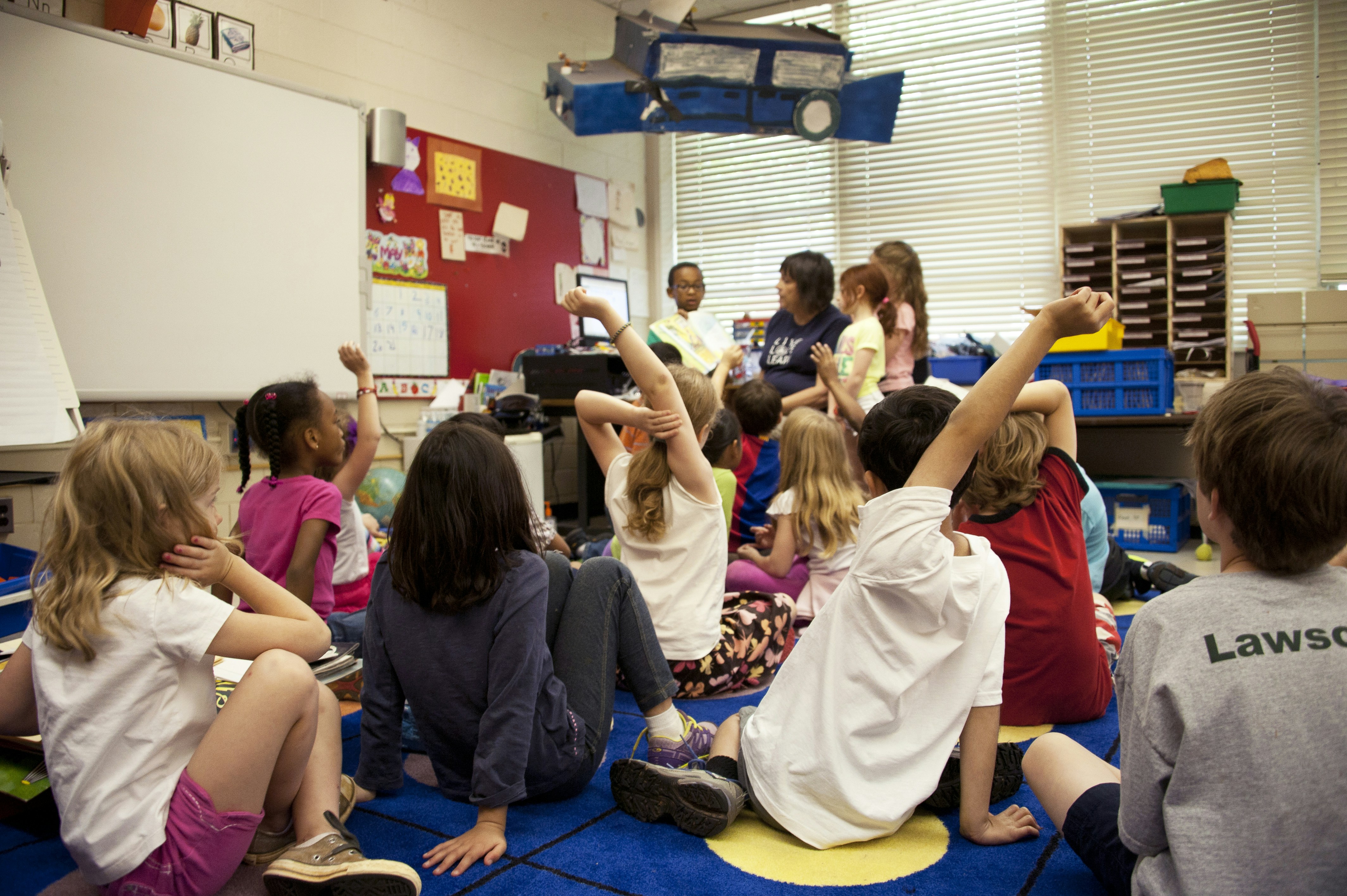 Children sitting on classroom floor raising hands.